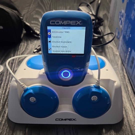 Compex FIT 5.0 sur sa station de charge, écran couleur affichant les programmes favoris et deux modules sans fil