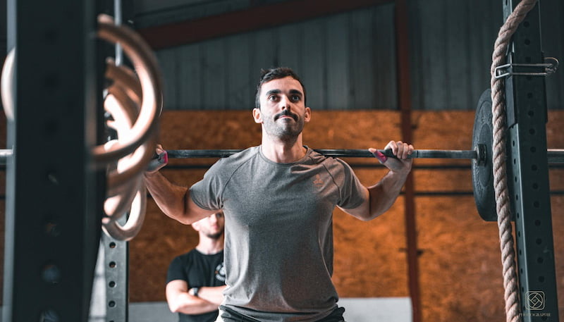 Gabin en back squat dans sa box de CrossFit, barre sur les épaules, coach en arrière-plan